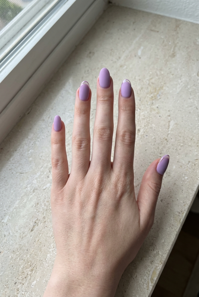 A hand positioned on a light stone countertop displaying a matte purple nail art idea with dusty purple polish and subtle tonal French tips on almond-shaped nails