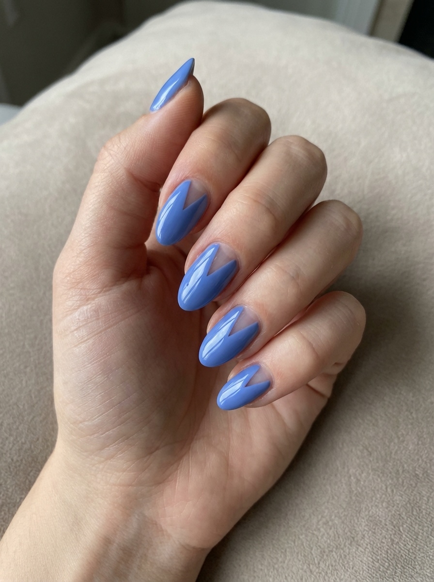 Hand photographed indoors against a soft beige background, highlighting a blue almond nail art idea with almond-shaped nails finished in glossy blue chevron-style tips.