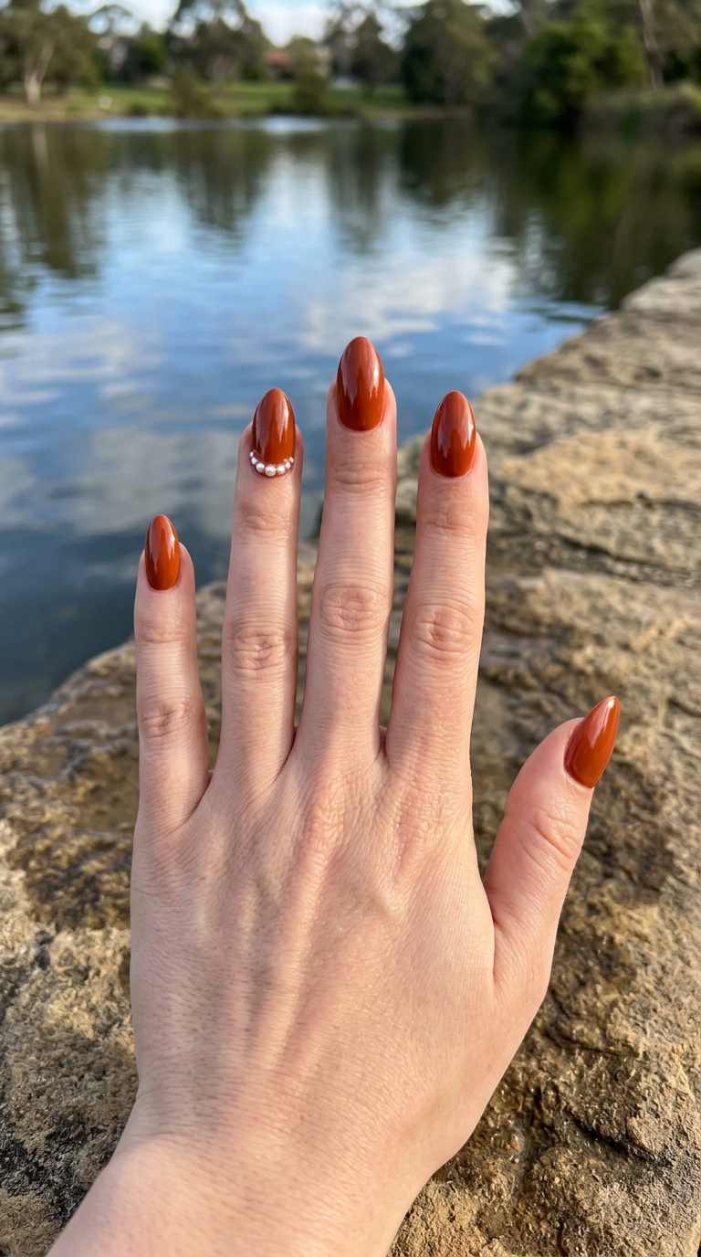 Fingers extended near a calm lake with rocks in the foreground, featuring an orange almond nail art idea with shiny rust-orange nails and a pearl-accented nail design