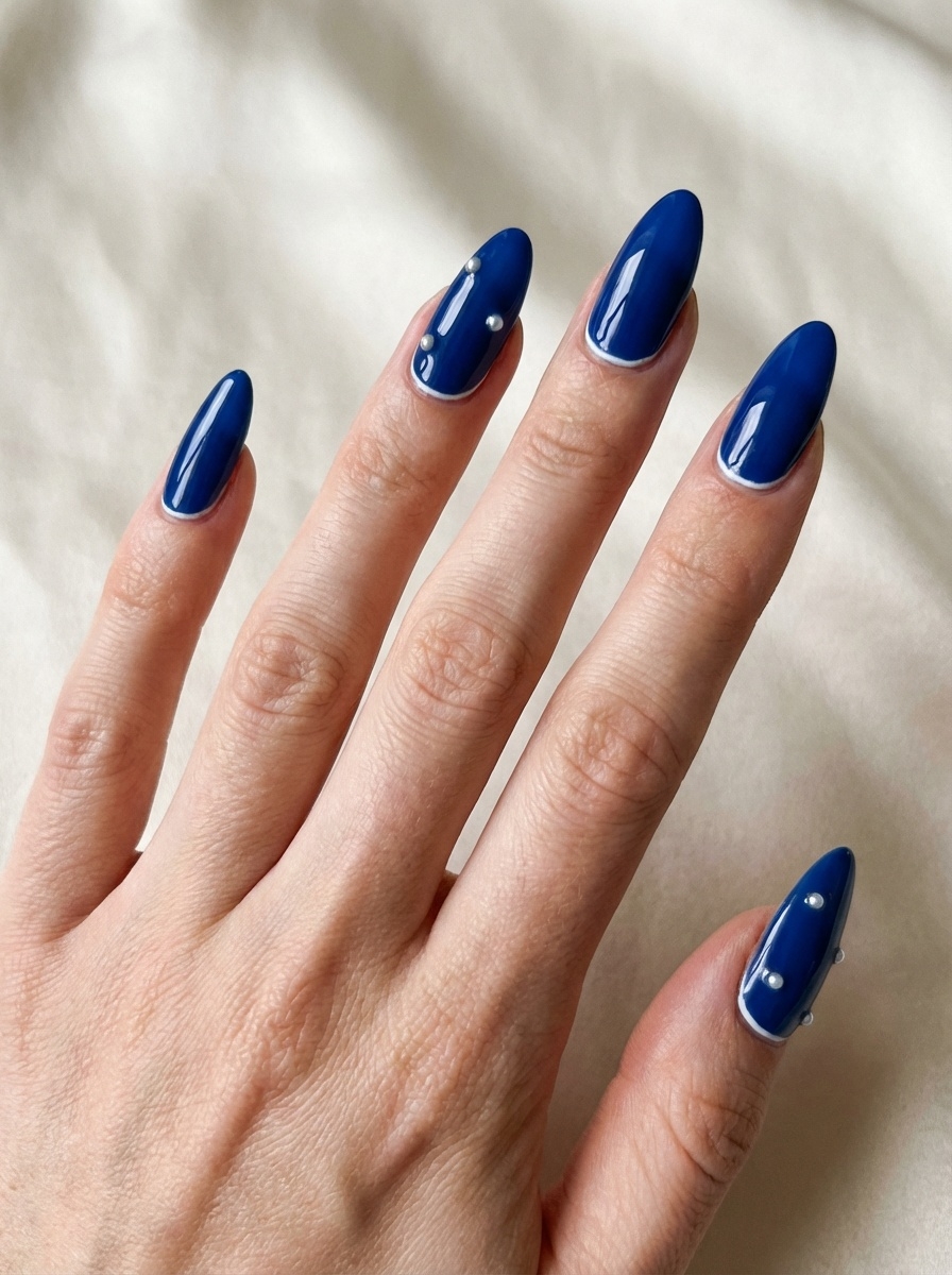 A close-up of a hand resting on soft fabric, showcasing a blue almond nail art idea with glossy cobalt nails detailed by small silver studs on select fingers.