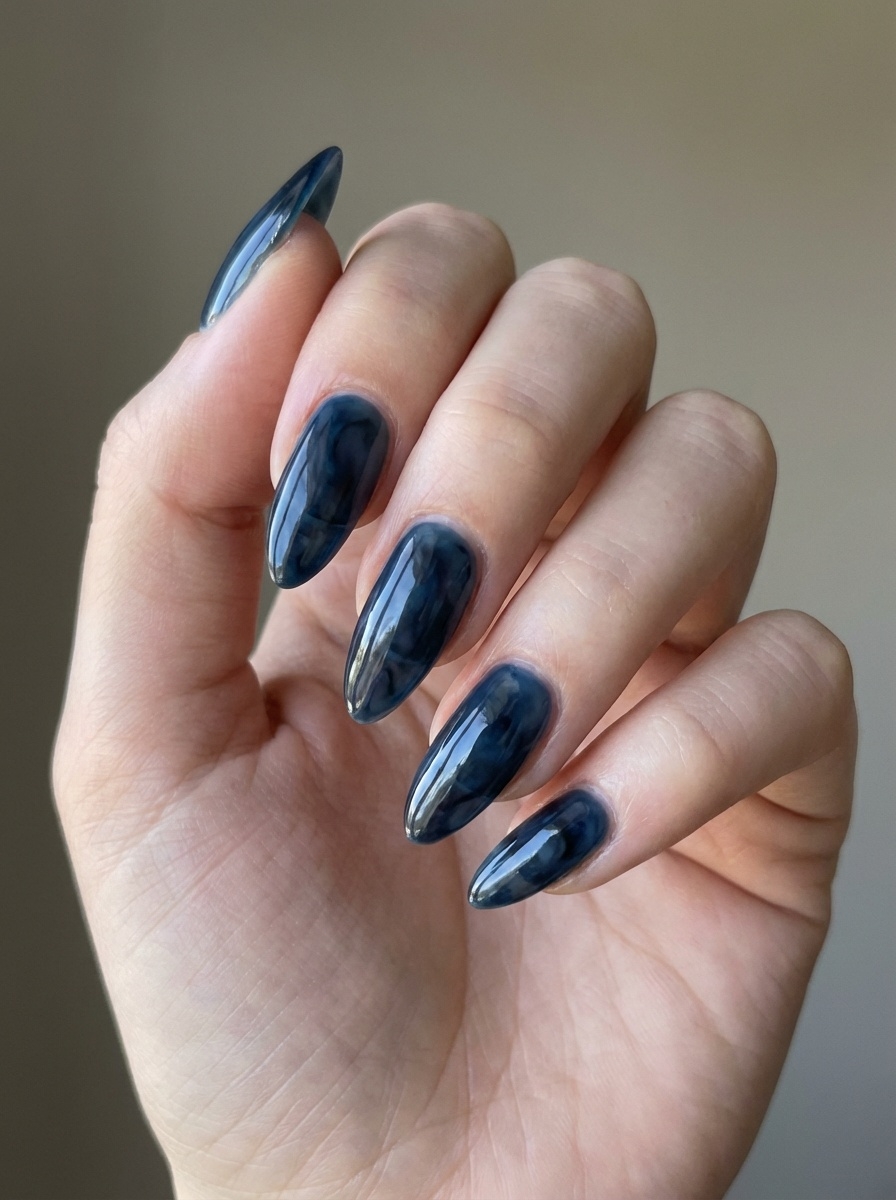 A softly lit hand photographed near a neutral background, featuring a blue almond nail art idea with reflective navy almond nails and a high-shine surface.