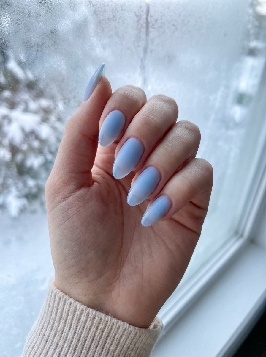 A hand near a frosted window wearing a blue almond nail art idea with matte light-blue almond nails, softly diffused lighting, and a cozy neutral sweater cuff.