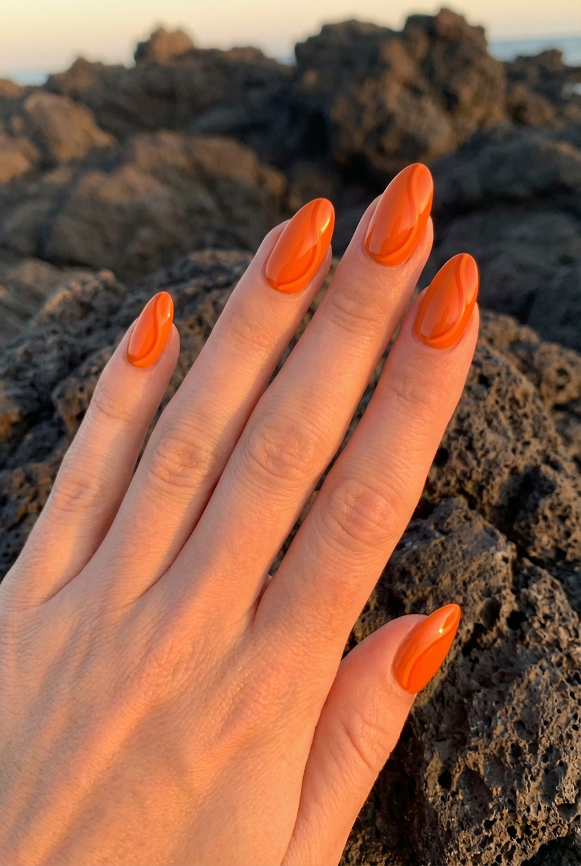 Close-up of a hand on seaside rocks during sunset, showcasing an orange almond nail art idea with vibrant glossy orange nails reflecting golden-hour lighting