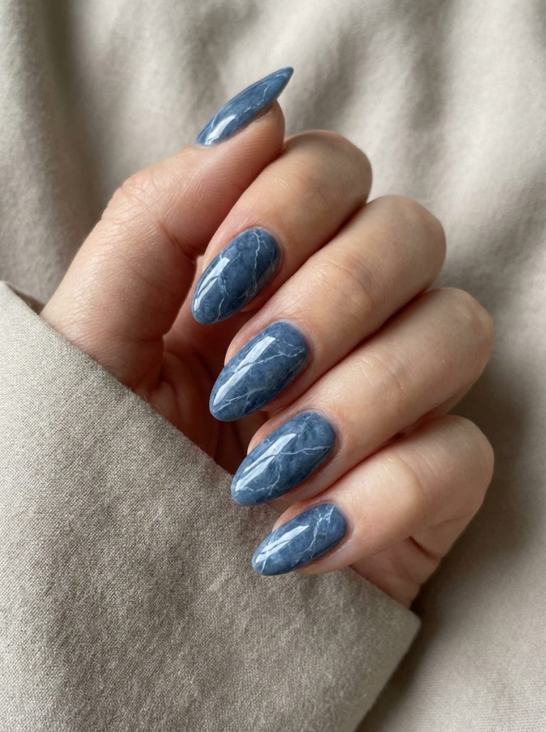 A hand with long, almond-shaped nails featuring a glossy blue marble design, displayed against a soft beige fabric background.
