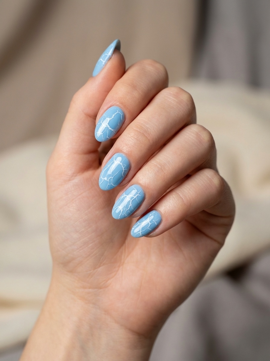 Close-up of a relaxed hand, displaying a blue almond nail art idea with cool blue almond nails finished with fine white crackle-style accents.