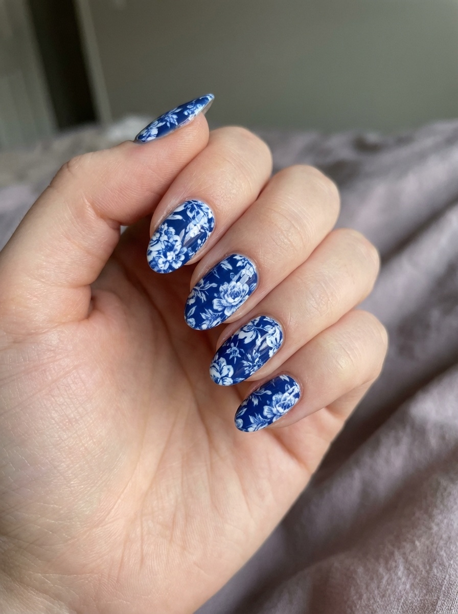 Close-up of a hand showing blue almond nails decorated with white floral patterns, photographed against soft neutral fabric.