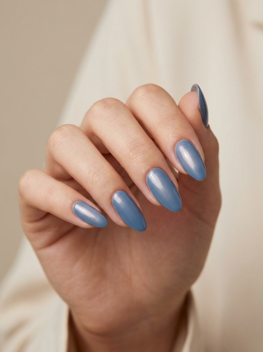 Hand resting against a light fabric backdrop, displaying a blue almond nail art idea with almond-shaped nails painted in a muted, sky-inspired blue gloss.