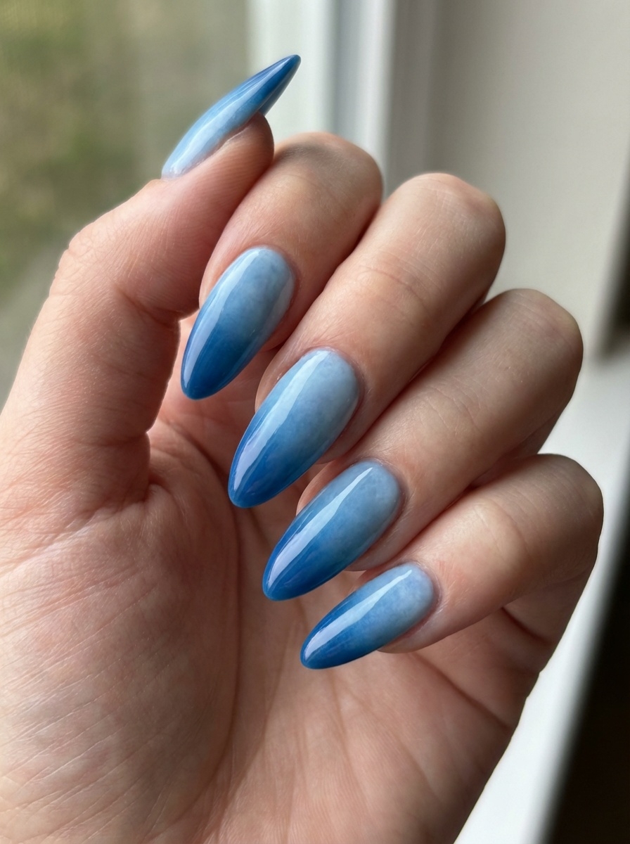 A hand angled toward a window, featuring a blue almond nail art idea with elongated almond nails finished in a refined blue ombré resembling ocean tones.