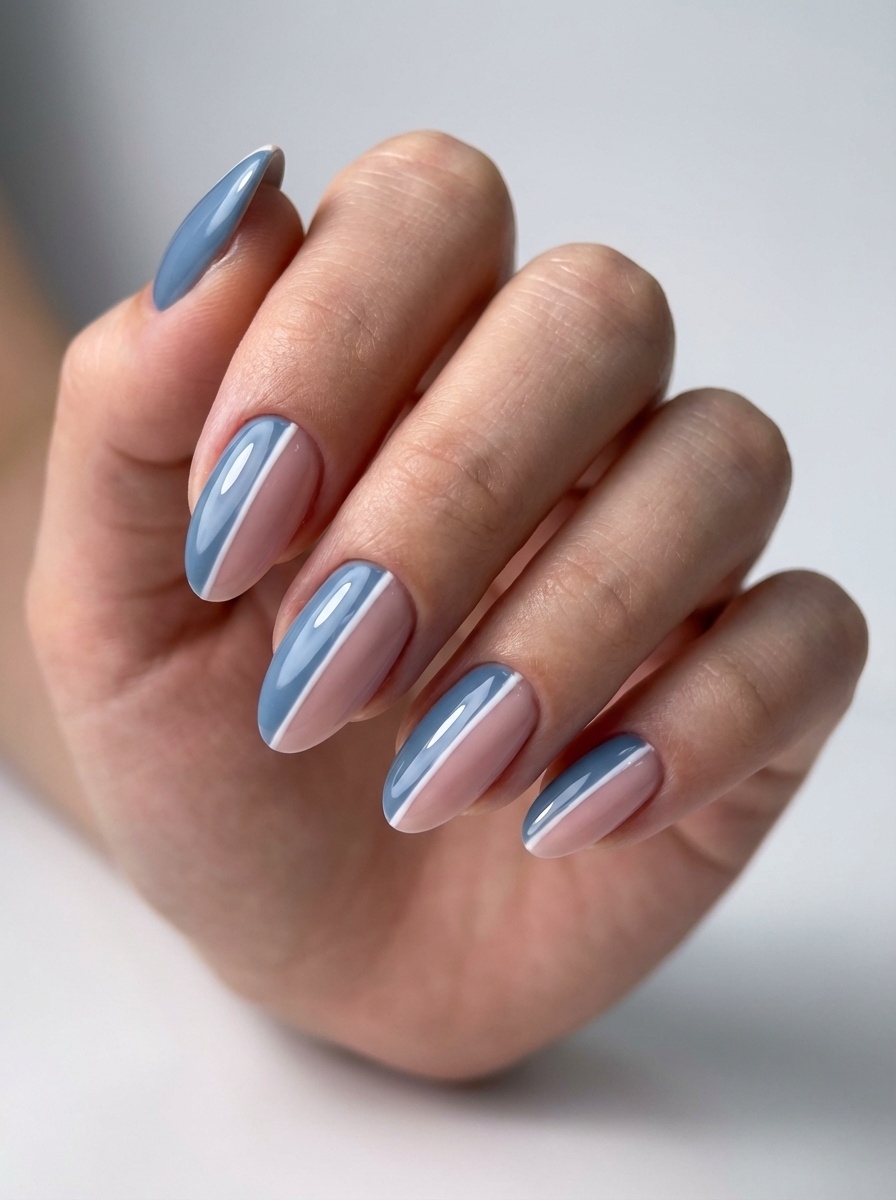 A close-up of a hand against a soft white background, featuring almond-shaped nails with a blue almond nail art idea combining blush pink centers and slate-blue side accents.