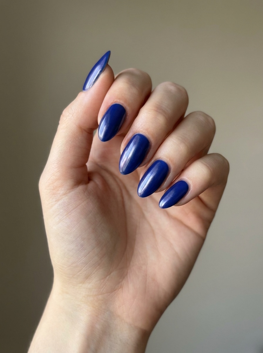 A close-up of a hand against a neutral wall, showcasing a blue almond nail art idea with glossy cobalt-blue almond-shaped nails and smooth reflective shine.