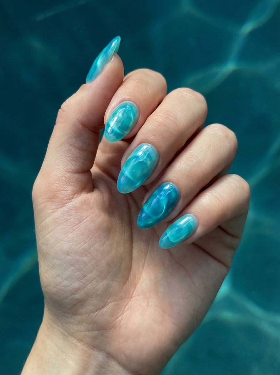 A hand held above rippling blue water, showcasing a blue almond nail art idea with translucent aqua-blue almond nails and a high-gloss, water-like finish.