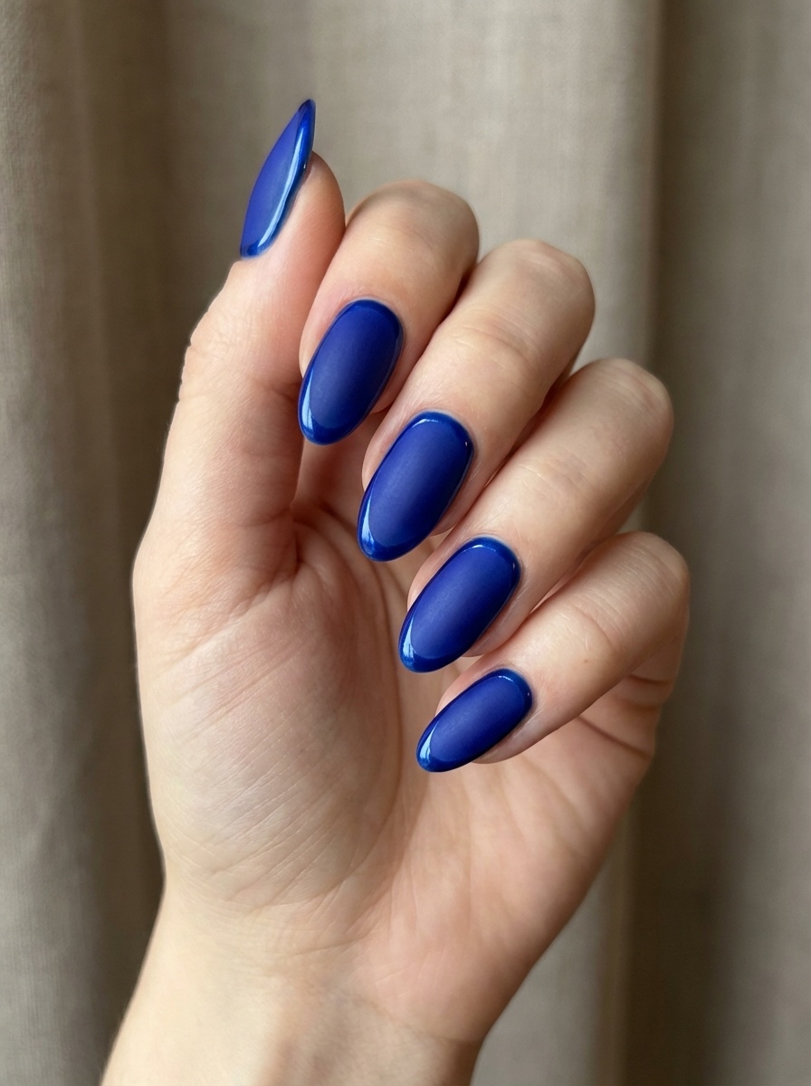 Hand posed near neutral fabric, featuring a blue almond nail art idea with deep royal blue almond nails finished in a softly reflective sheen.