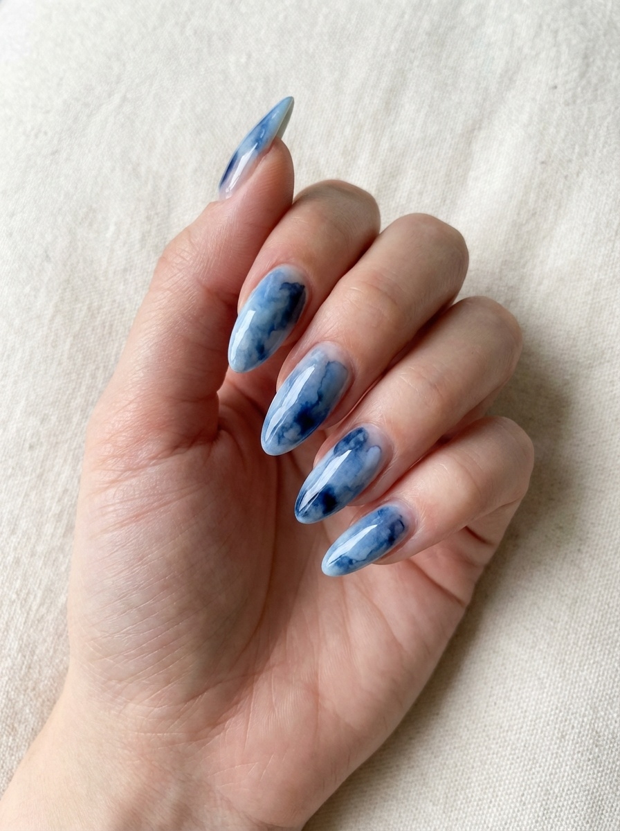 Close-up of a hand against beige textile, highlighting a blue almond nail art idea with marble-effect blue almond nails blending light and dark tones.