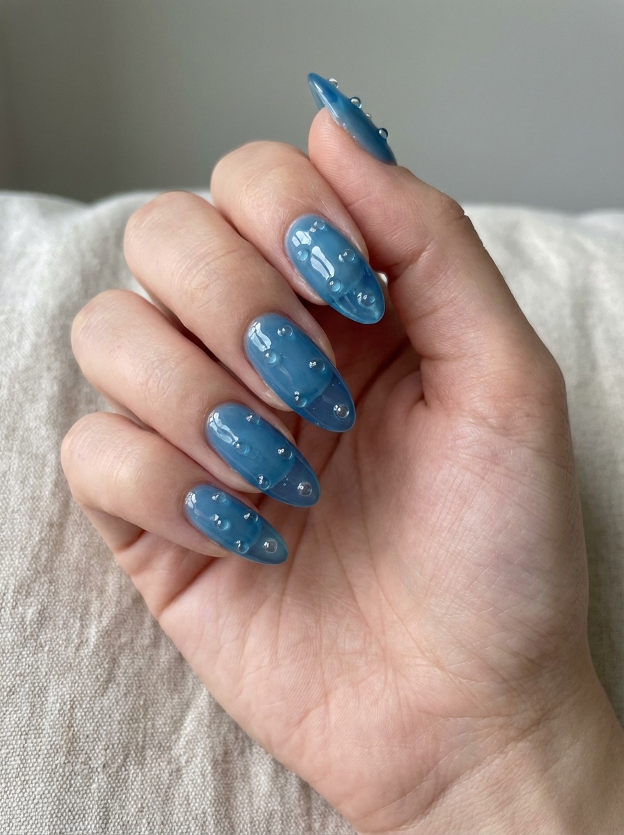 Close-up of a hand resting on fabric, featuring a blue almond nail art idea with sheer blue jelly almond nails accented by raised clear droplets.