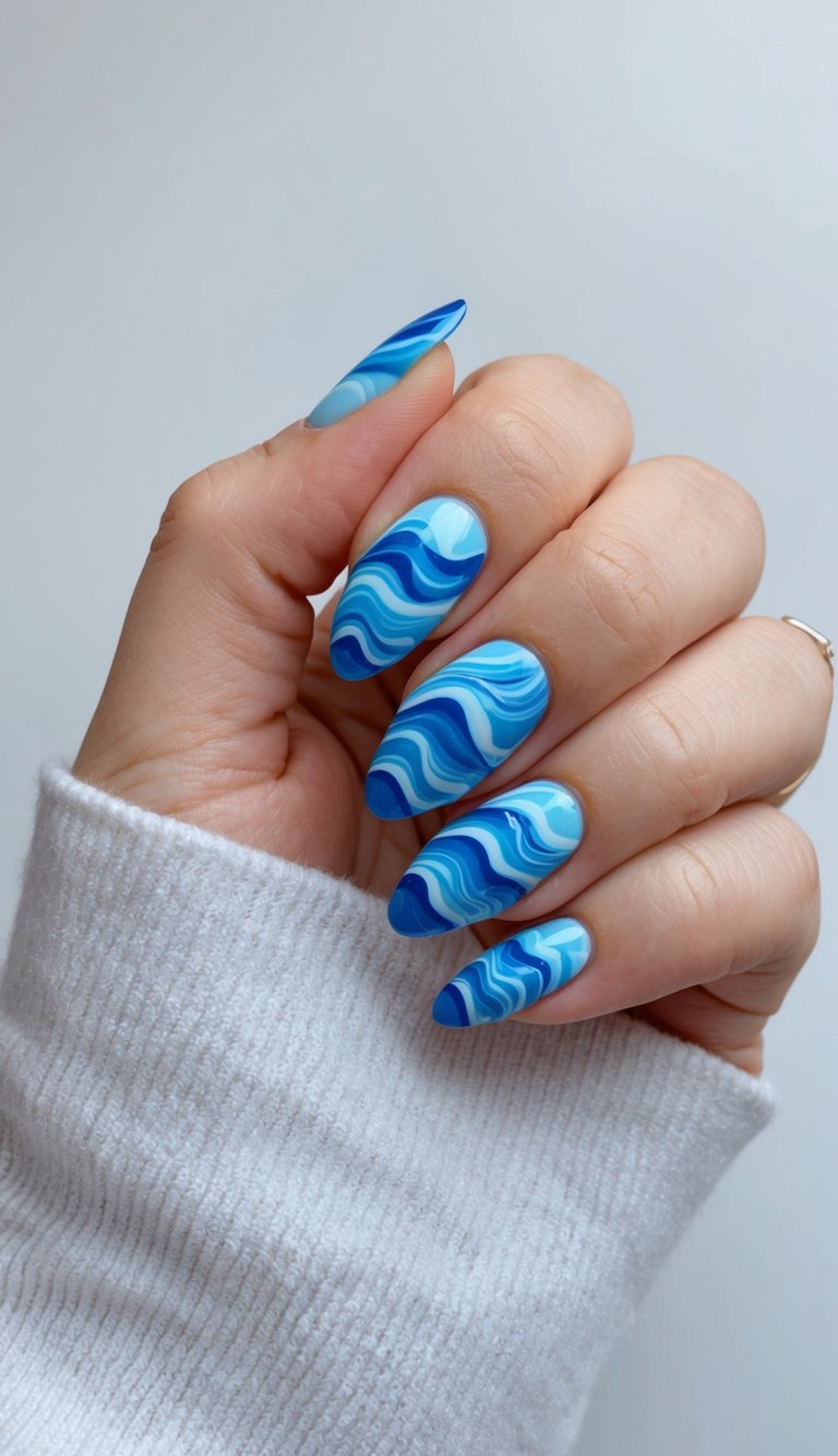 A manicured hand partially covered by a white knit sleeve, featuring a blue almond nail art idea with almond-shaped nails decorated in flowing blue wave patterns and glossy layered finishes.