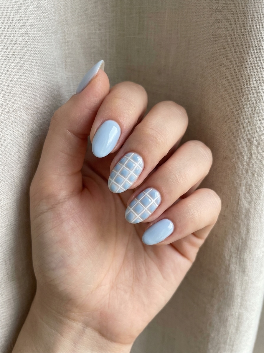 Hand resting against neutral fabric, featuring pale blue almond nails with a glossy finish and a delicate white grid accent on two nails.