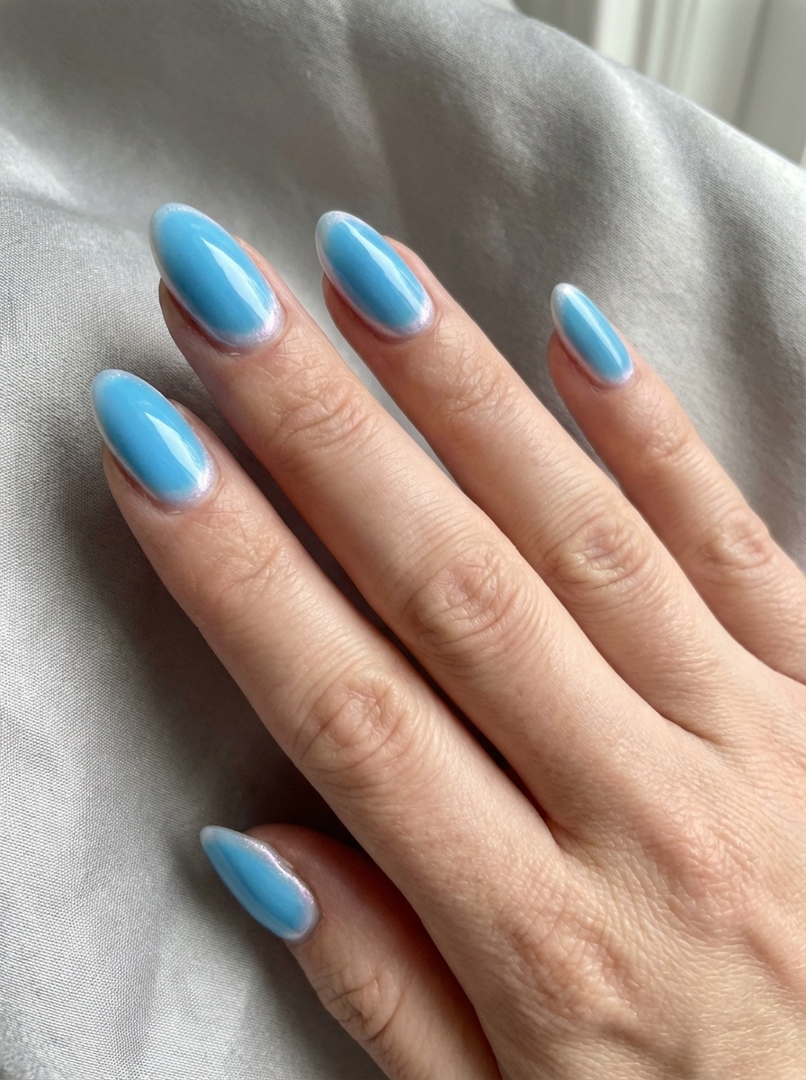 A hand resting on neutral fabric showcases almond-shaped nails in pastel blue, finished with a subtle white sheen outline along the edges for a refined blue almond nail style idea.