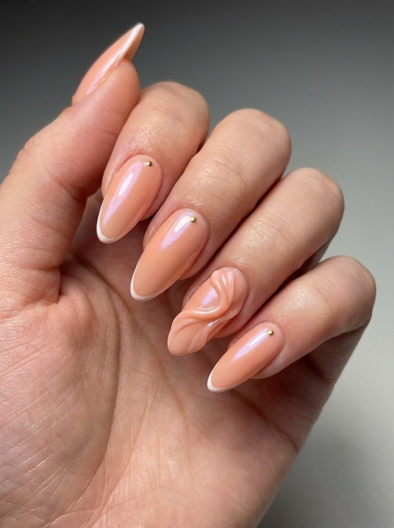 A hand with long almond-shaped nails painted in a glossy peach color, featuring subtle white tips and a small gold stud on each nail. The ring finger showcases an intricate textured design with a wavy pattern.