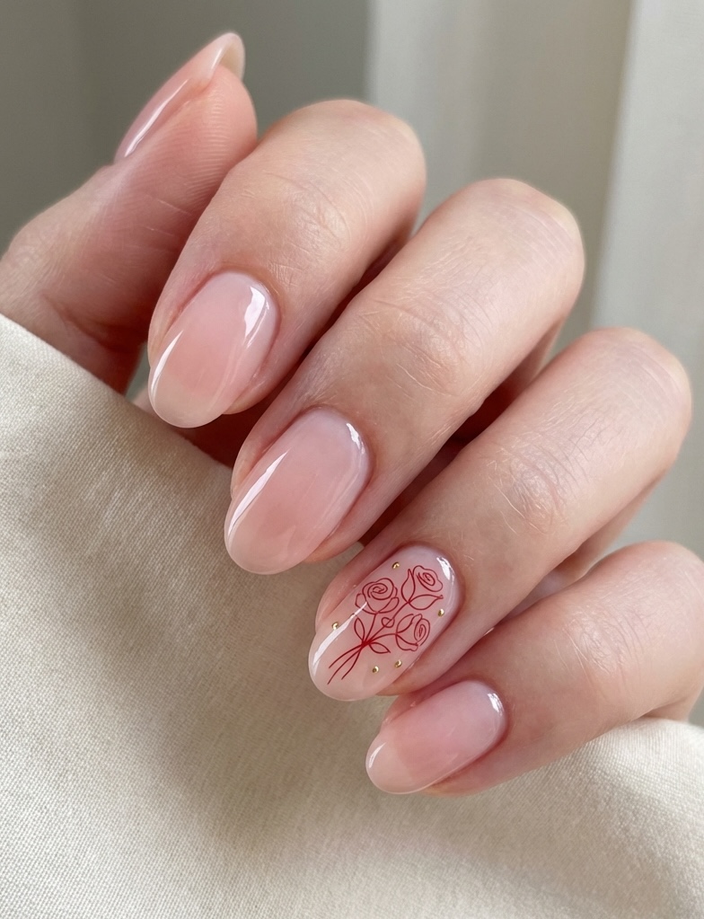 Angled view of fingers resting against beige fabric, revealing glossy blush nails and one accent nail detailed with red rose drawings and fine gold embellishments for valentine's nail art ideas.