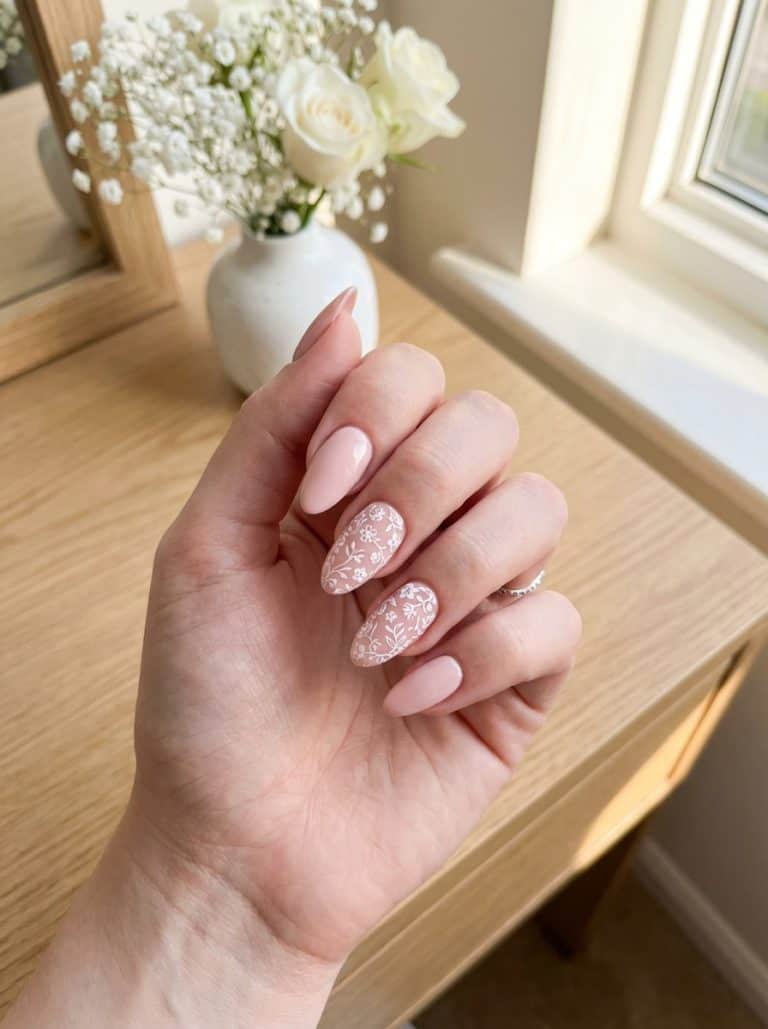 A hand with long almond-shaped nails painted in a soft pink color, featuring intricate white floral designs on two nails. The background includes a wooden surface and a white vase with white roses and baby's breath flowers placed beside a window.