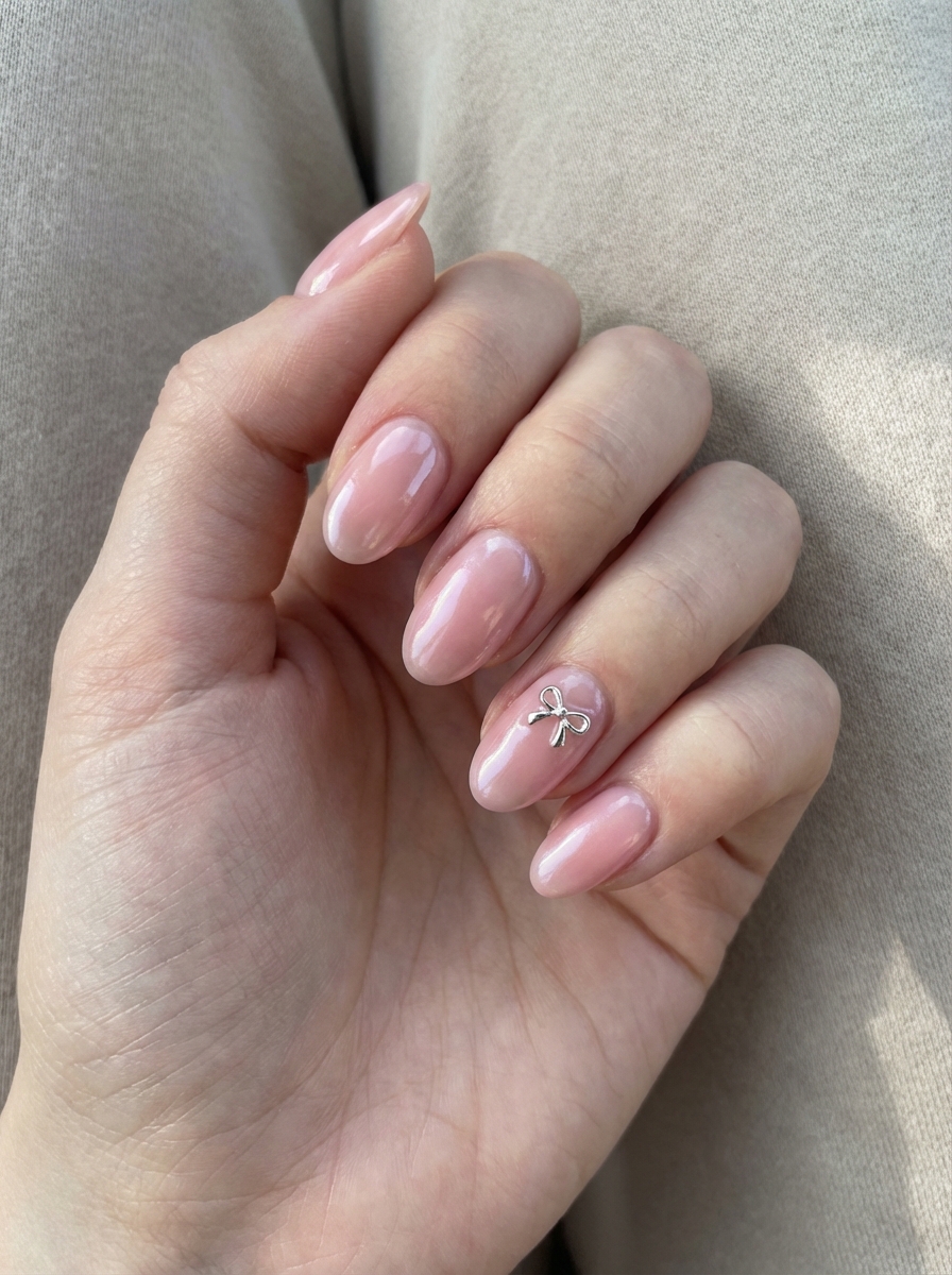 Hand with shiny soft pink nails and a small silver bow charm on the ring finger—an adorable and sweet prom nail art idea.