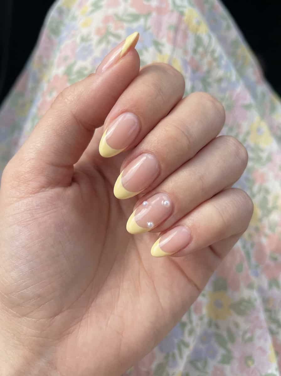 A hand featuring a yellow nail art idea with creamy yellow French tips, set on glossy nude nails and decorated with small pearl accents on the ring and middle fingers.
