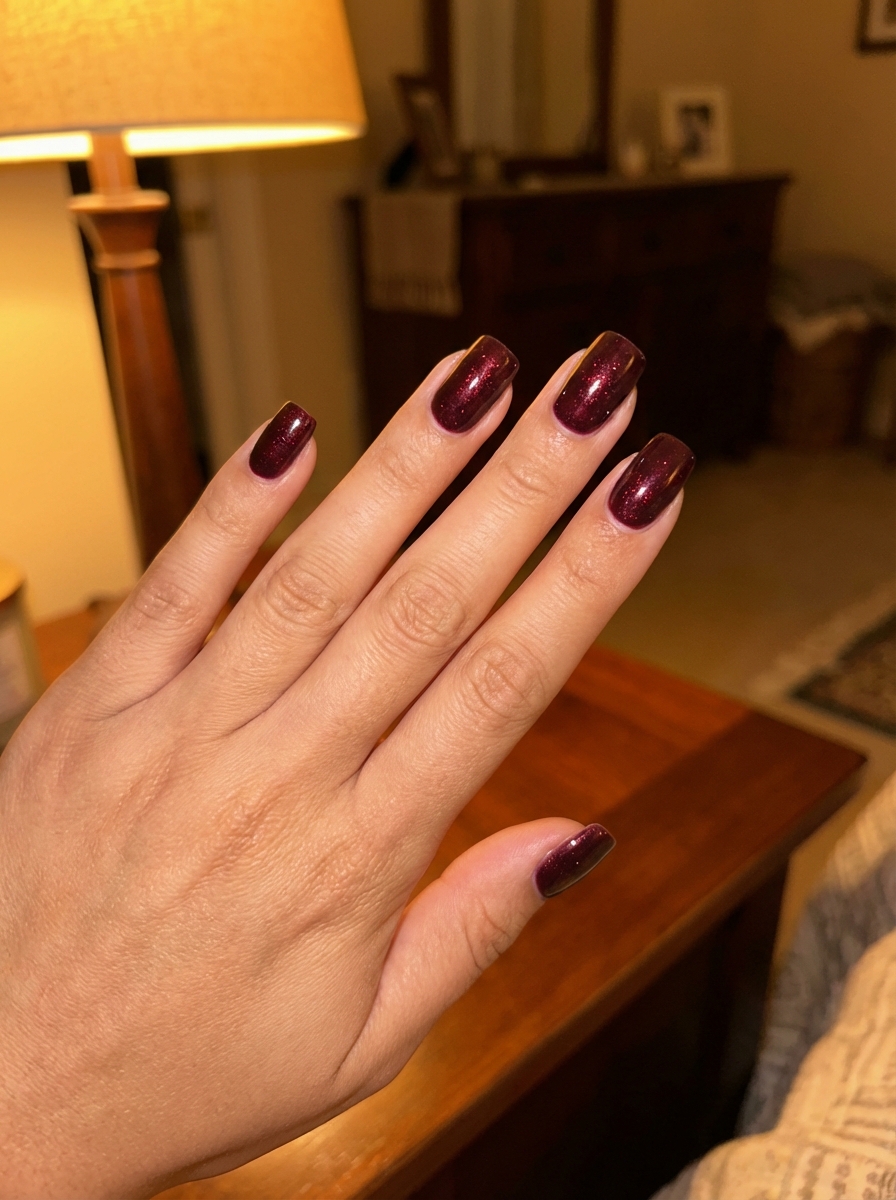 Close view of a manicured hand inside a softly lit bedroom featuring a bridesmaid nail art idea with dark burgundy shimmer polish and squared nail shape