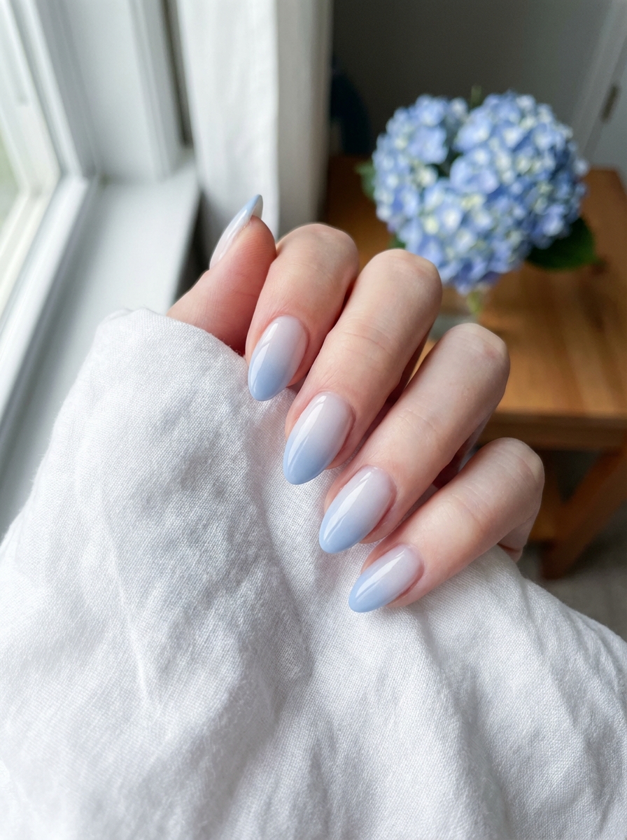 Close-up of a manicured hand showing a bridesmaid nail art idea with milky white to pastel blue ombré nails photographed beside a hydrangea bouquet on a wooden table