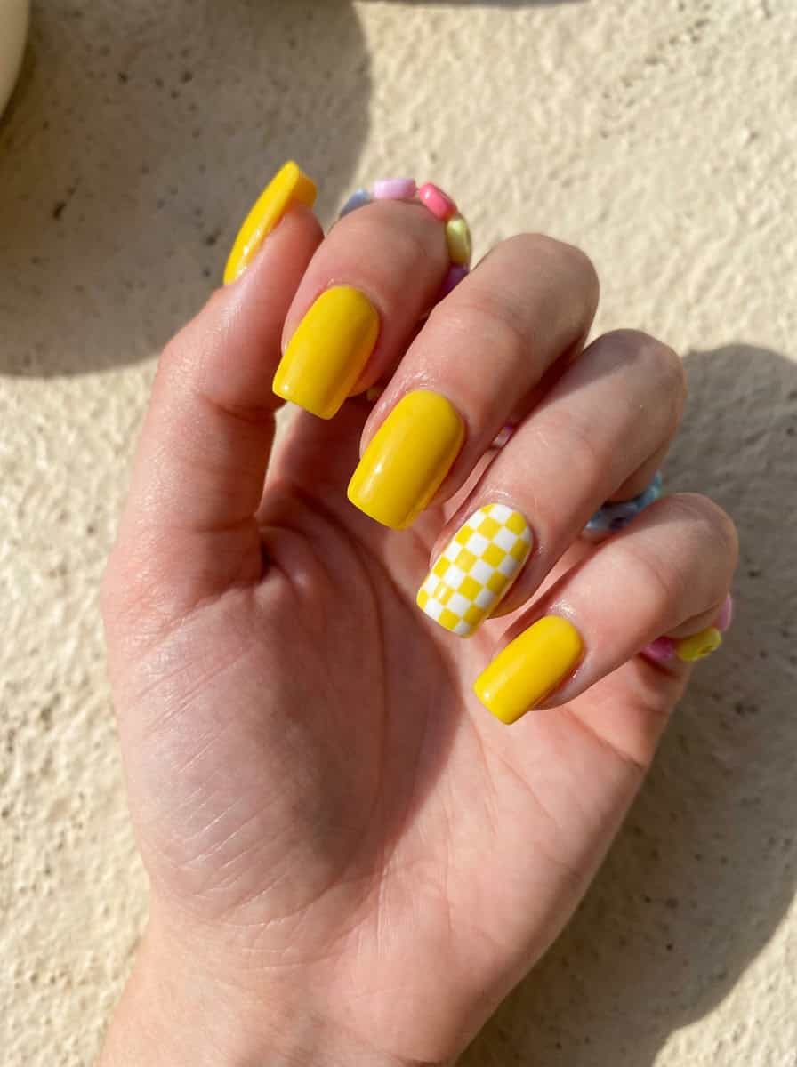 A hand showcasing a yellow nail art idea with four glossy yellow nails and one nail decorated in a yellow-and-white checkerboard design, bathed in natural light.