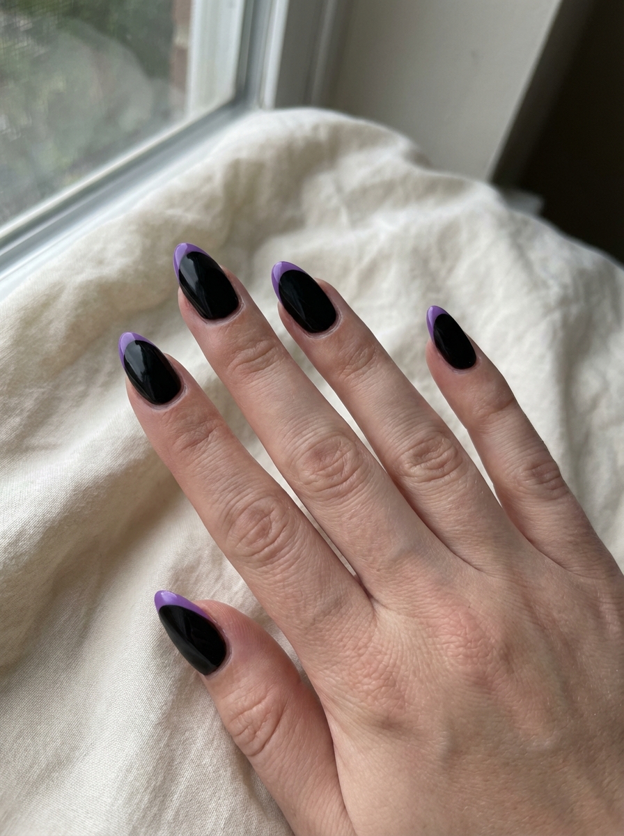 Fingers stretched across soft neutral cloth reveal jet-black almond nails topped with subtle lilac tips, illustrating a minimalist purple and black nail art idea photographed near a window.