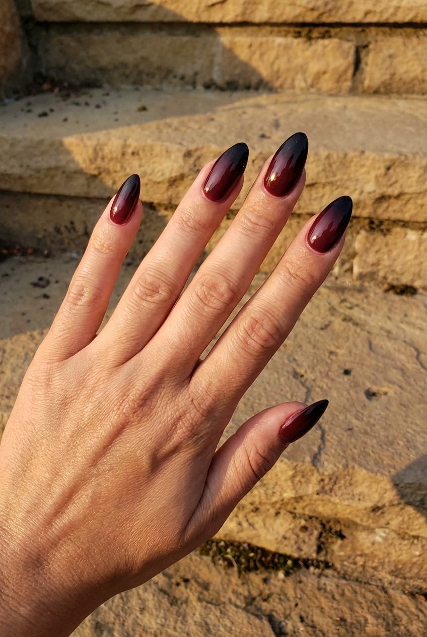 Against sunlit stone steps, a hand reveals a maroon nail art idea with long almond nails featuring a smooth gradient shifting from wine maroon near the base to glossy black tips.