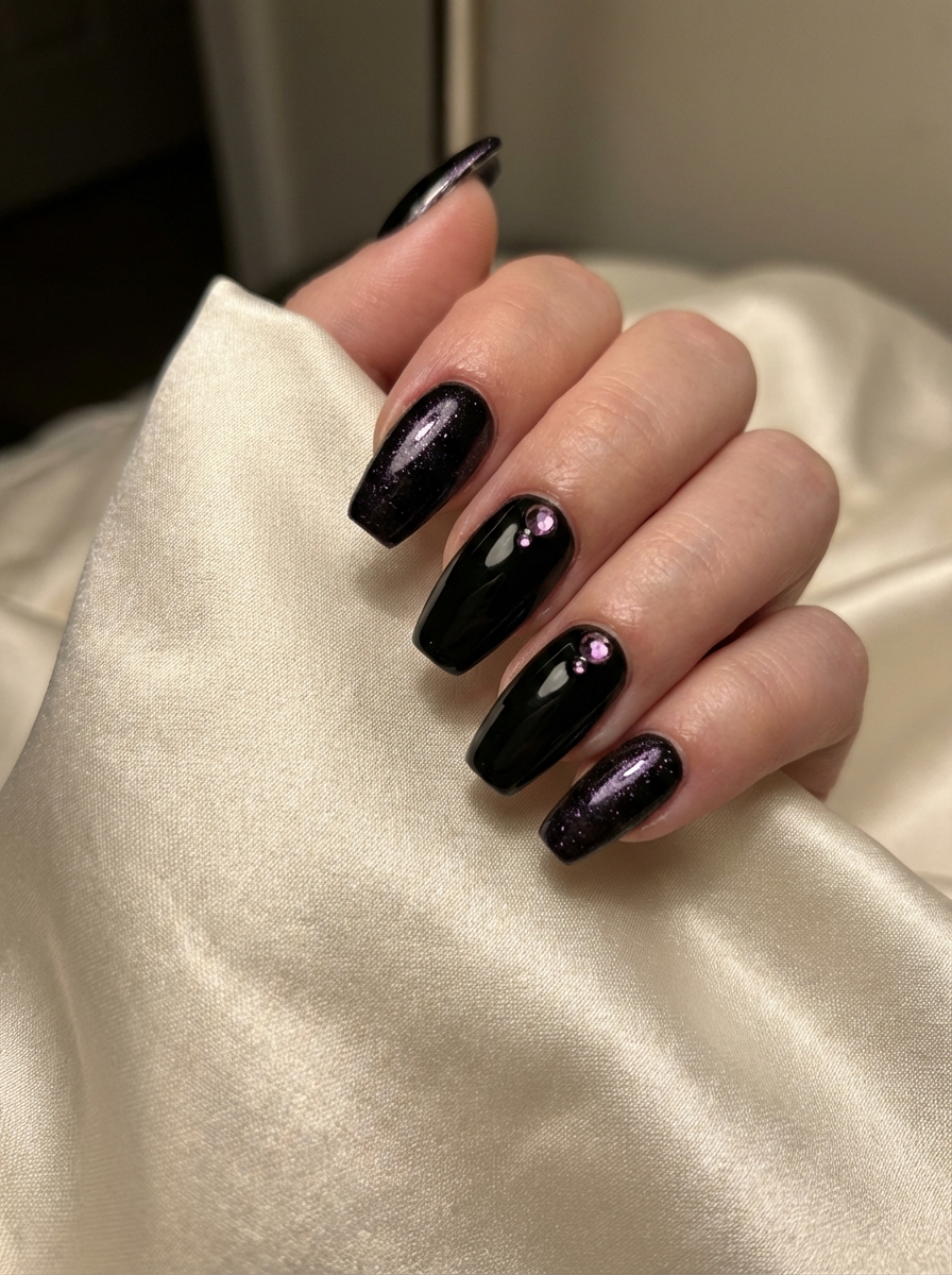 Under warm indoor light, a hand holds silky fabric while long black nails appear beside glittery purple accent nails adorned with small crystals in a purple and black nail art idea.