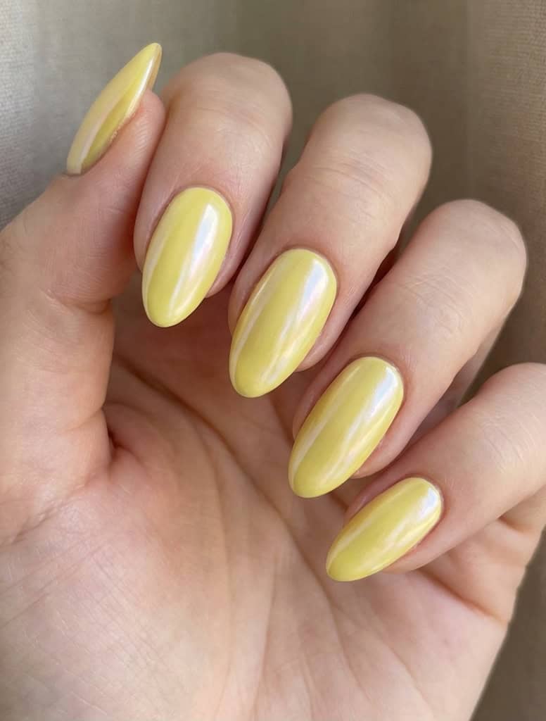 A hand showing a yellow nail art idea with long oval nails coated in creamy pastel yellow polish and subtle pearl shimmer, captured in soft daylight by a beige background.