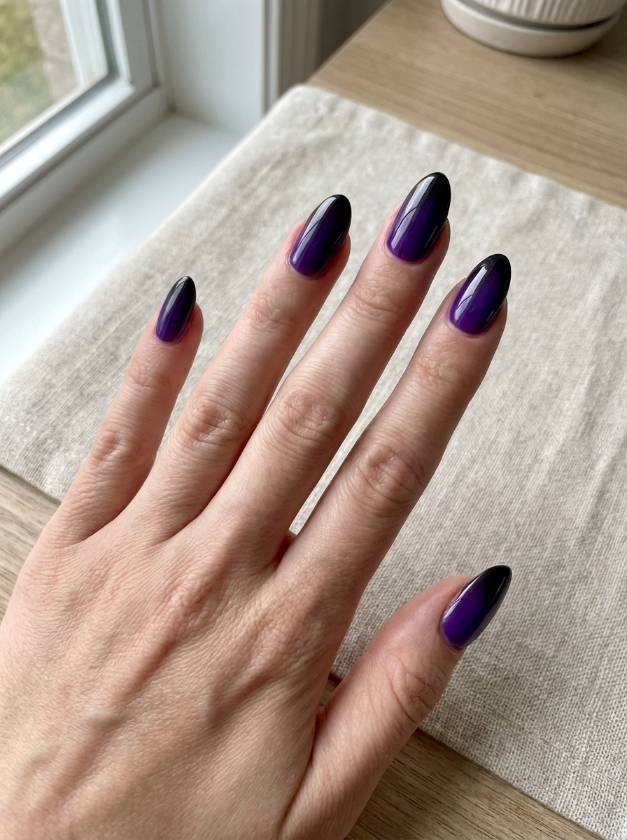Resting above a light linen surface near a window, fingers reveal long almond nails with a violet-to-black gradient finish, highlighting a purple and black nail art idea.