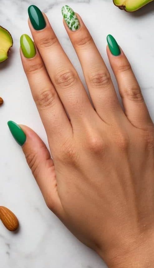 A marble surface with scattered avocados and almond nails in various shades of green, creating a fresh and trendy nail art display