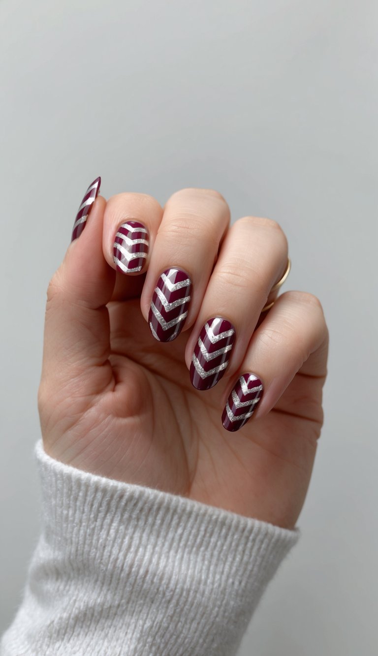 Hand slightly curled with a thin ring visible, showcasing a maroon nail art idea with glossy wine maroon nails decorated with layered silver chevron stripe patterns.