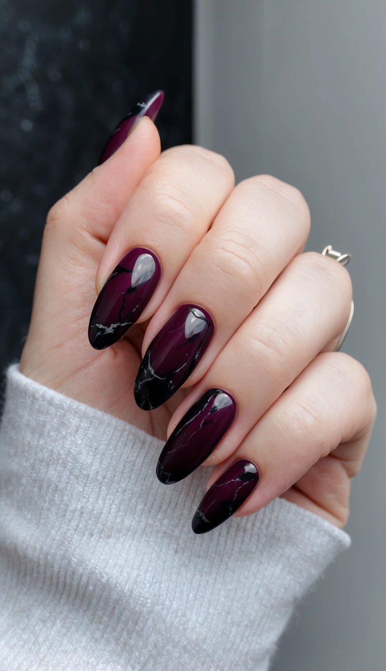 Hand raised near a neutral background featuring a maroon nail art idea with almond-shaped nails in deep maroon fading into black marble tips with subtle white veining.