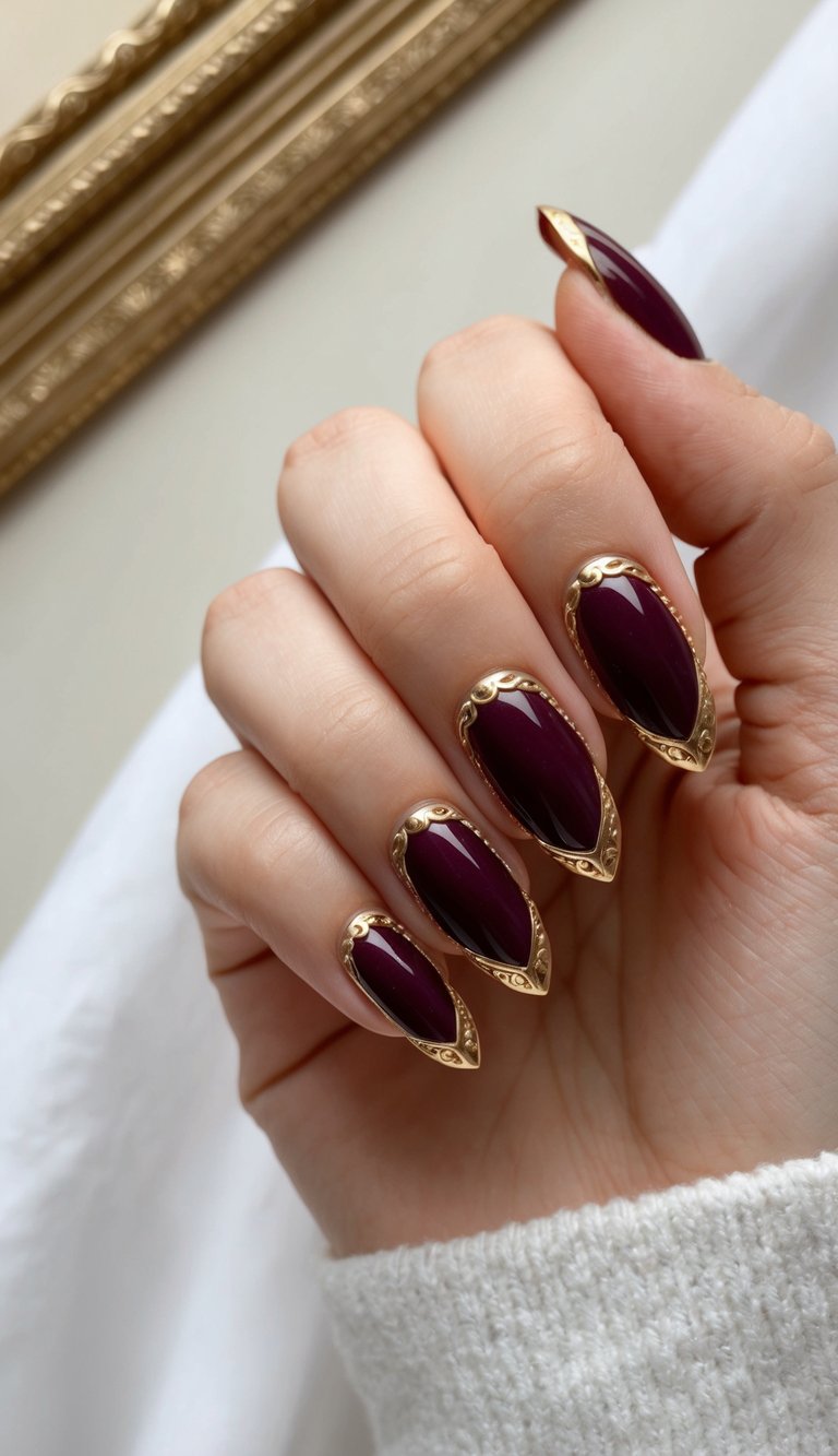 Close-up of a manicured hand showing a maroon nail art idea with glossy burgundy nails surrounded by ornate metallic gold borders against a soft indoor background.