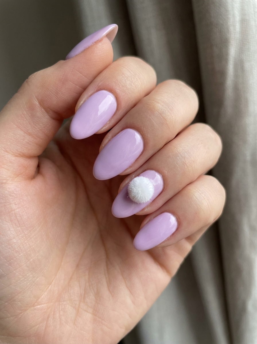 Hand with oval lilac nails showing a playful Easter nail art idea featuring a fuzzy white pom-pom on the ring finger as a bunny tail.