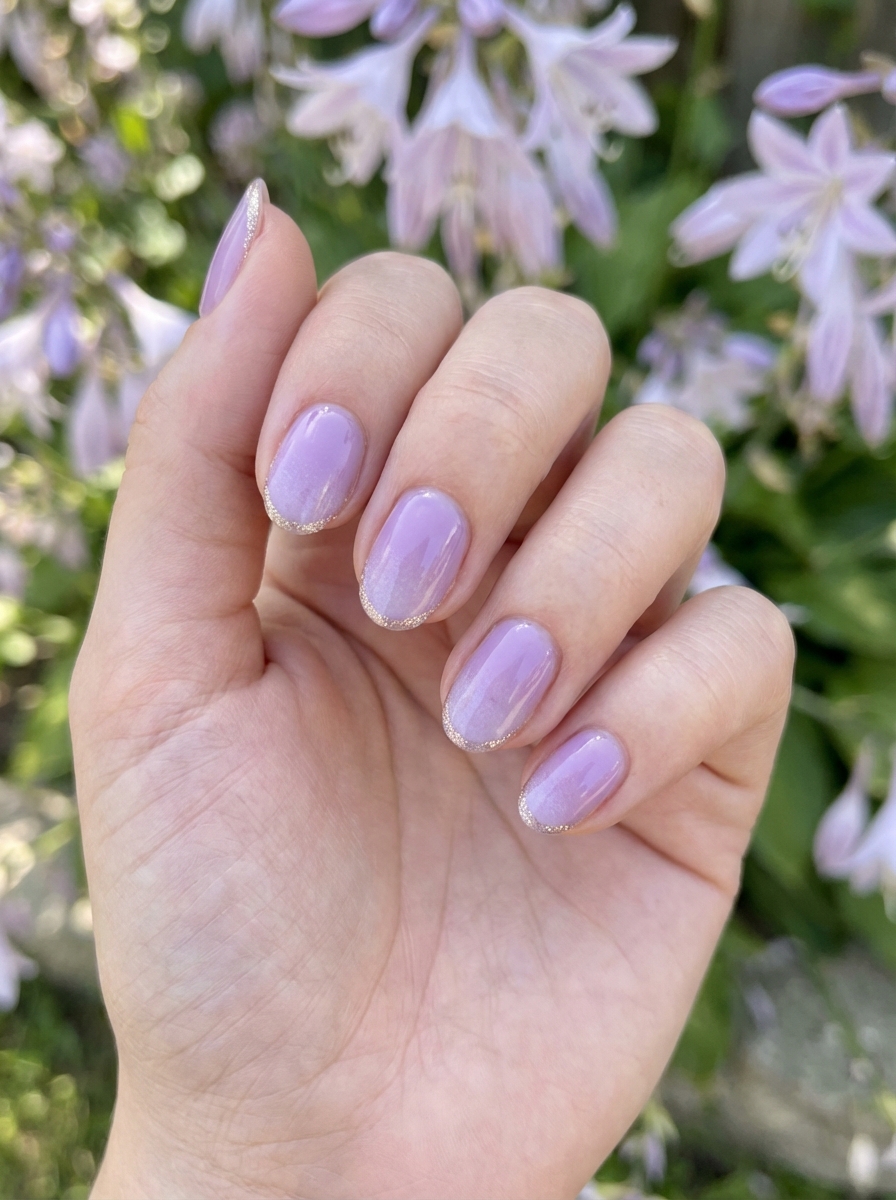 Short nails painted in a sheer lavender shade with fine gold glitter tips, showcasing a delicate lavender nail art idea near outdoor florals.