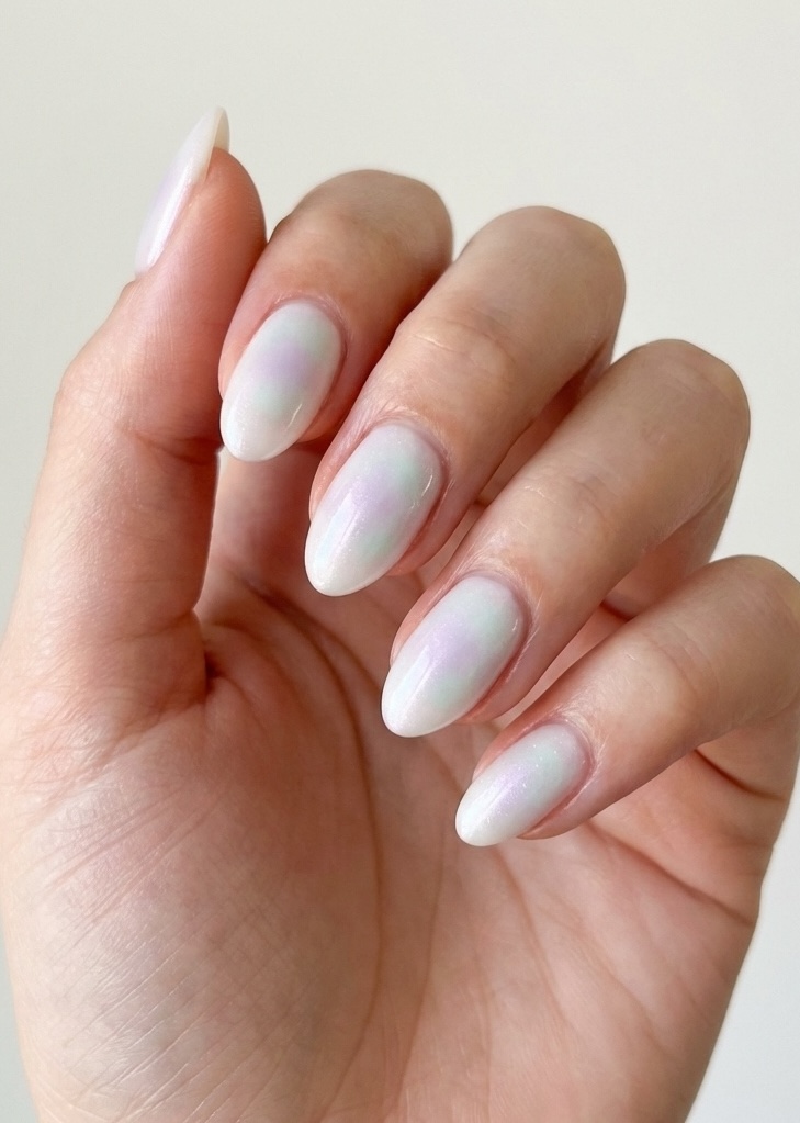 Lifestyle-style image of a hand featuring an aesthetic nail art idea with pearly white almond nails, iridescent pastel reflections, and a clean minimalist setting.