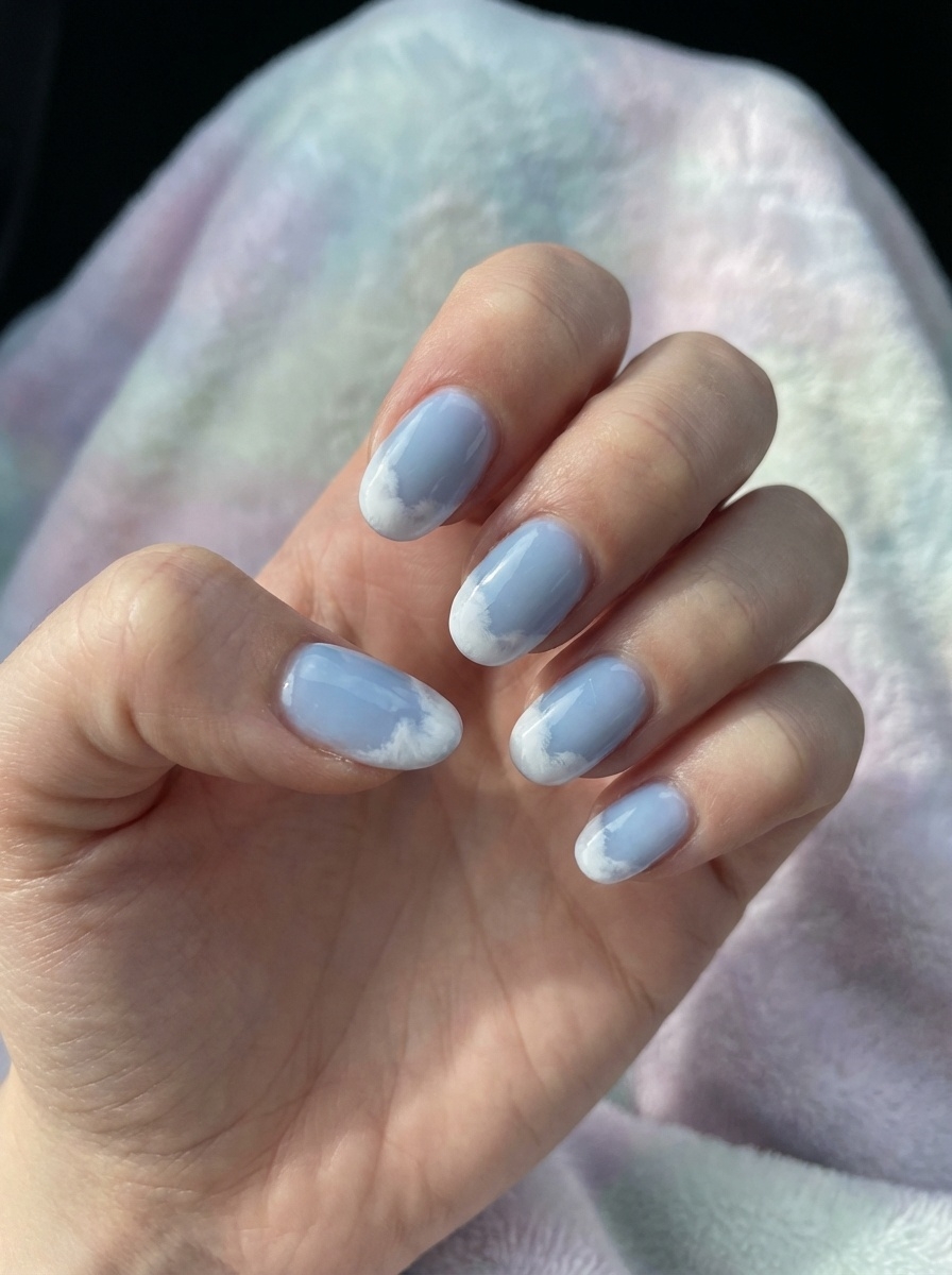 Close-up of fingers resting on soft pastel fabric, featuring an aesthetic nail art idea with light blue nails, cloudy white tips, and a smooth glossy finish.