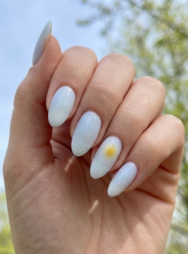 Angled outdoor beauty shot highlighting an aesthetic nail art idea with almond-shaped milky white nails, cool-toned sheen, and a delicate yellow sun design on one nail.