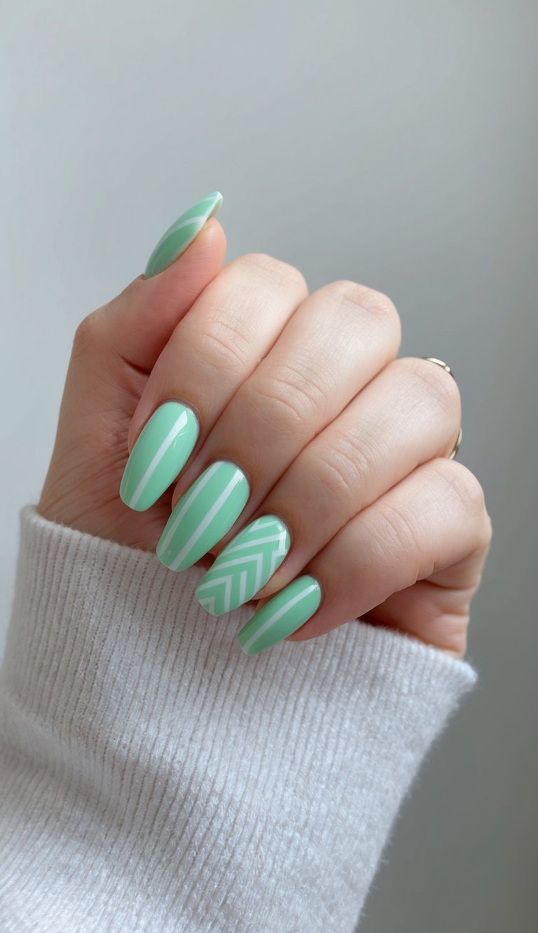 A hand wrapped in a cozy sweater sleeve features pastel mint green nails with crisp white stripes and a chevron accent, showcasing a refined mint green nail art idea.