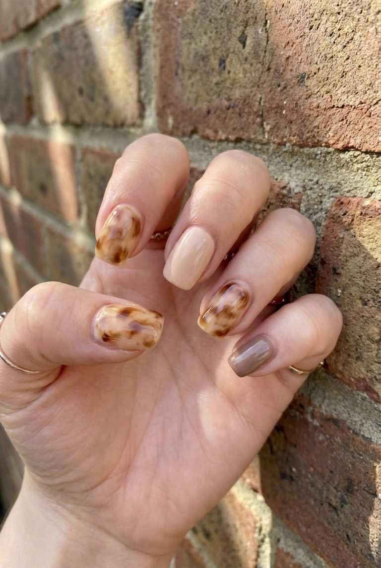 Close-up of a hand with manicured nails featuring a mix of tortoiseshell and nude designs against a brick wall background.