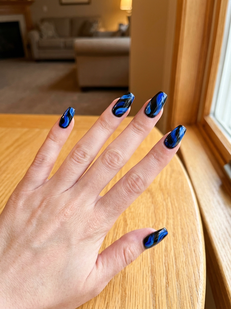 Fingers extended over a wooden tabletop displaying a black and blue nail art idea with striking sapphire-blue marble swirls layered over glossy black nails.