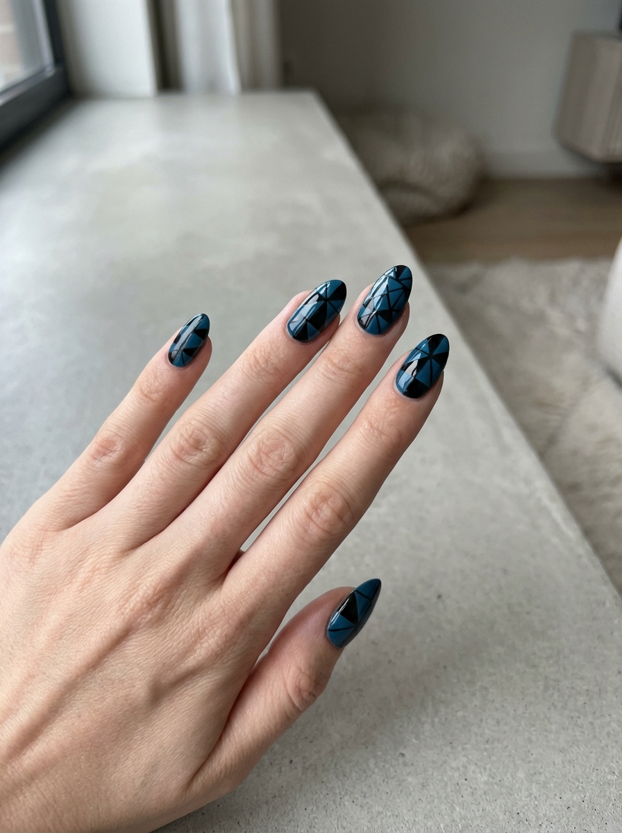 Hand resting on a light gray countertop near a window displaying a black and blue nail art idea with deep teal-blue nails featuring glossy black geometric triangle patterns.