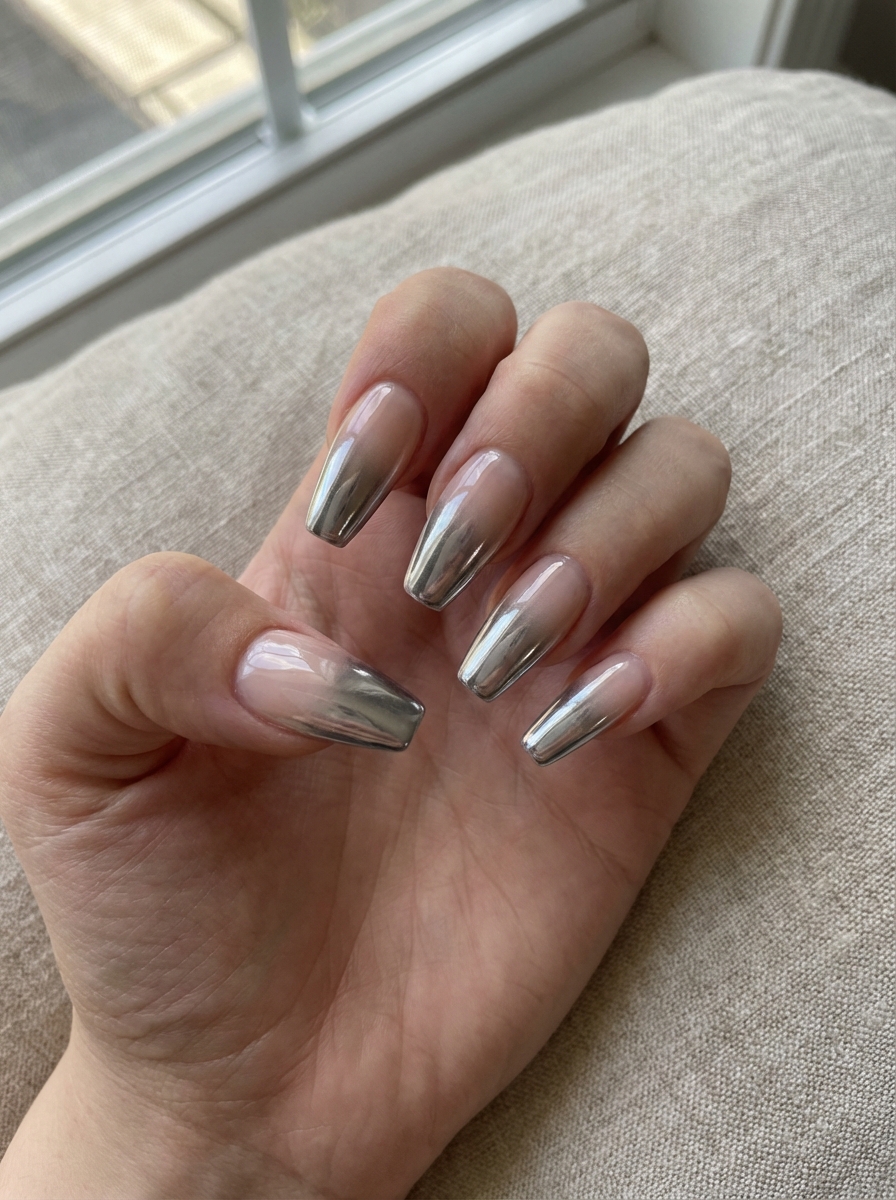 A hand with long nails featuring a chrome ombre design, with a gradient from natural to metallic silver tips.