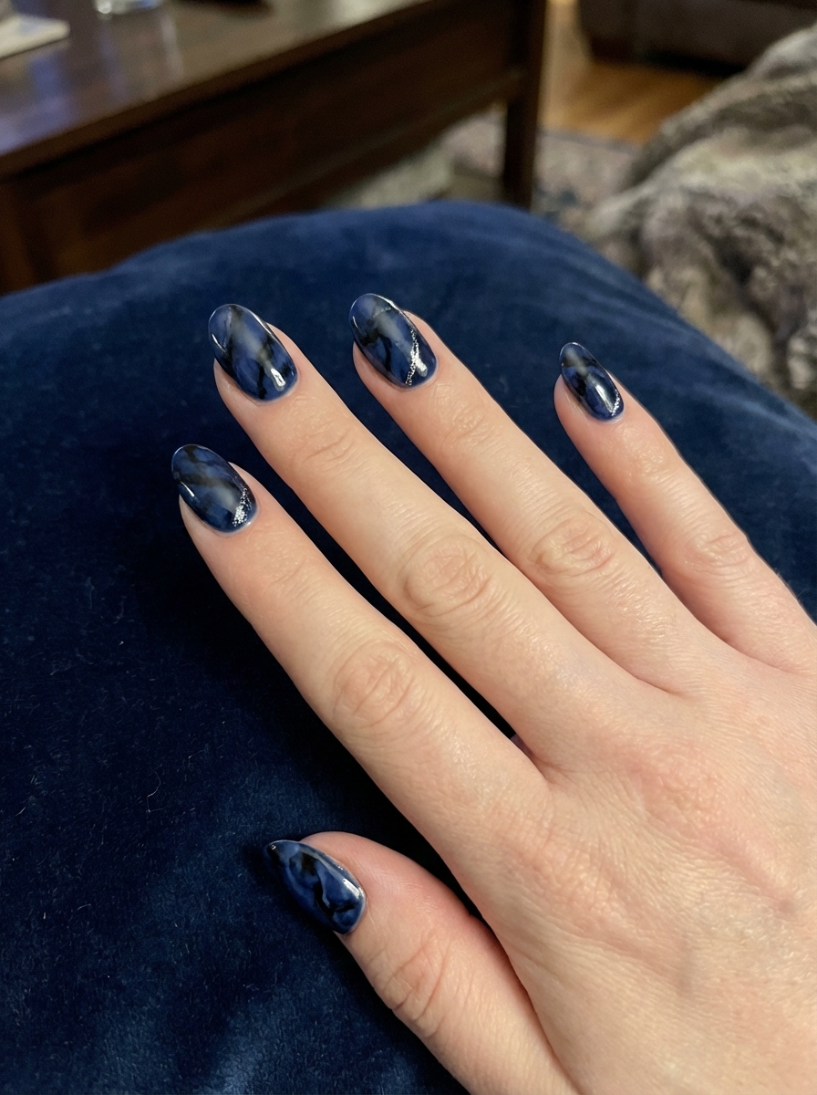 Almond-shaped manicure photographed over dark navy fabric featuring a black and blue nail art idea with swirling marble patterns in deep blue and black and thin silver streak accents.