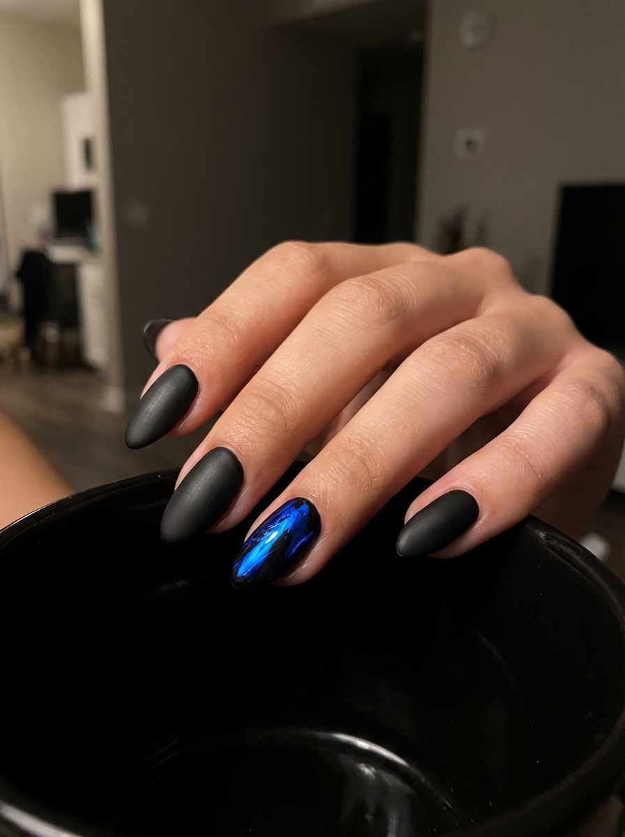 Fingers curved over a black bowl displaying a black and blue nail art idea with matte black almond-shaped nails contrasted by one bright metallic blue accent nail.